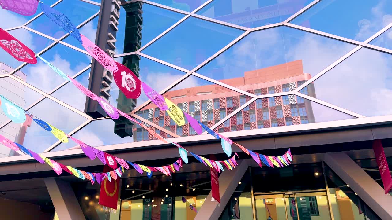 Vibrant paper flags flutter in the wind above a modern outdoor plaza in Singapore, captured with upward camera movement and bright daylight