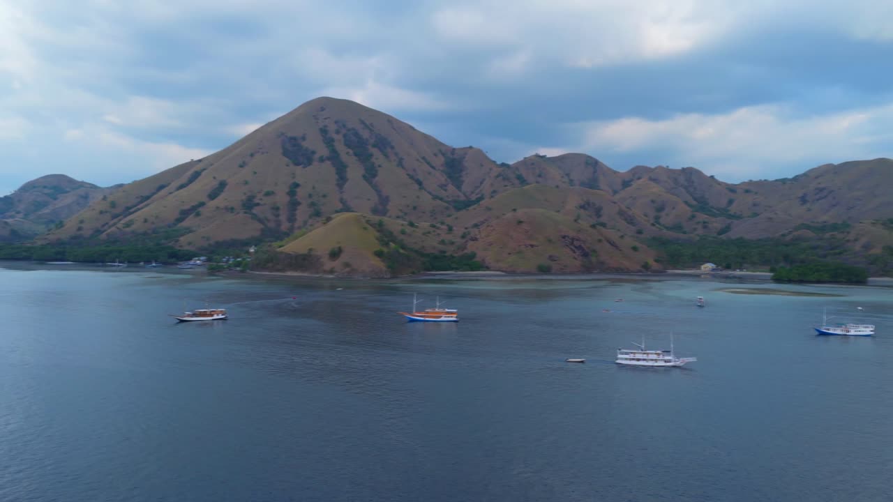 Aerial wide shot of traditional boats anchored in a calm bay surrounded by golden hills under a partly cloudy sky in Komodo