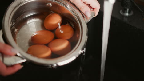 close up of woman hand holding metal bowl under running water in sink, bowl filled with brown eggs while faucet flows, scene captures moment of turning bowl to drain water during preparation