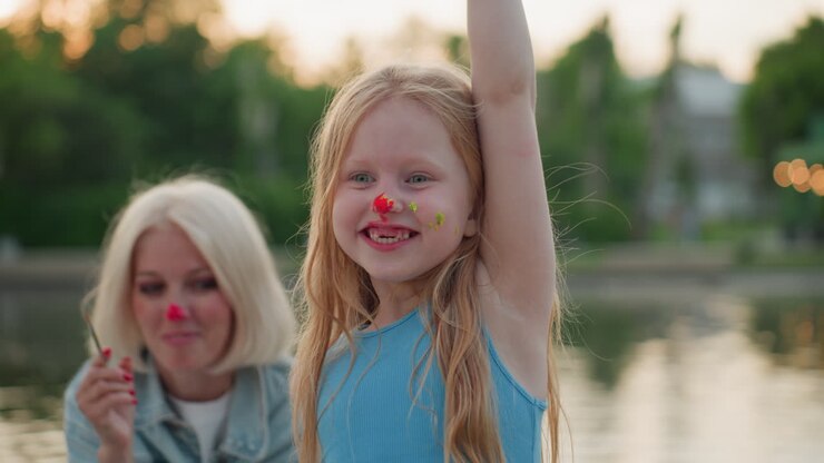 Ritratto di mamma sorridente e figlia giocosa che ballano in riva al lago al tramonto con volti dipinti e pennelli, una sessione d'arte gioiosa, puntini colorati su guance e naso, una calda serata, divertimento creativo all'aperto.