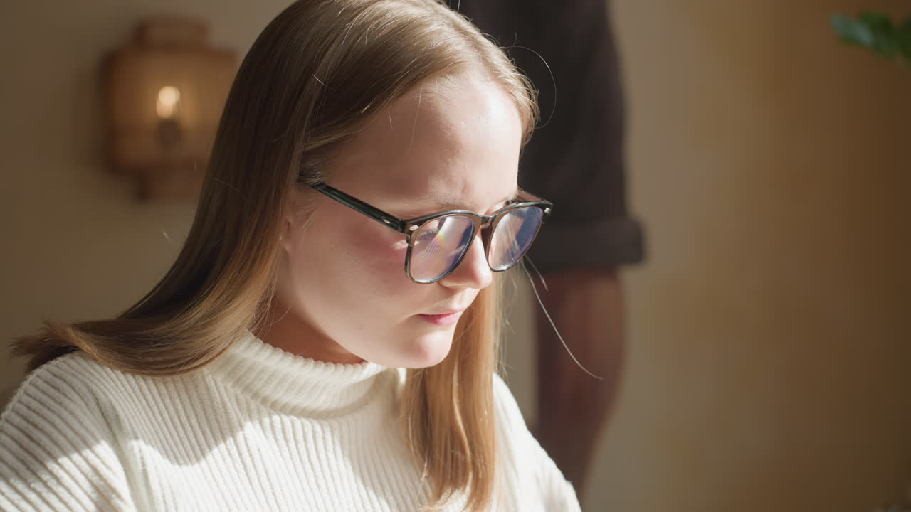 Thoughtful lady in white sweater reading book near window as sunlight softly illuminates her face, blur background showing wall art, indoor plant, and coffee cup on wooden table