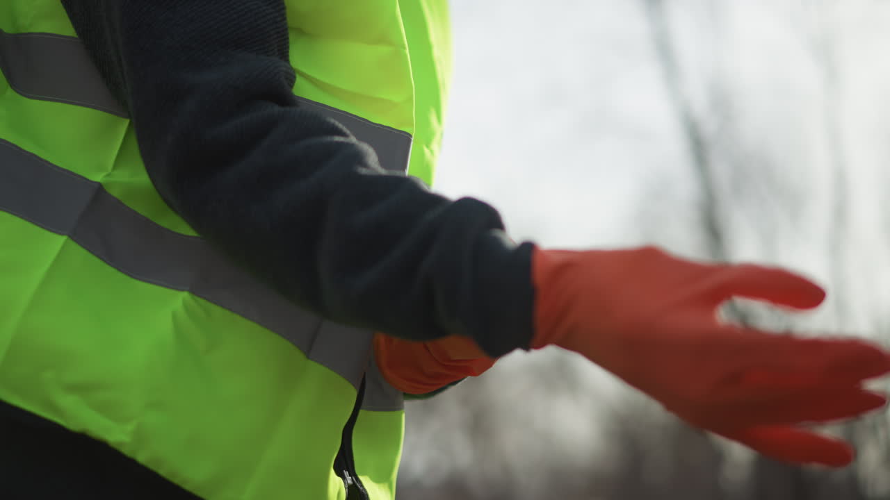 Worker in dark sweater and high visibility vest putting on bright orange protective gloves outdoors, preparing for safety work in construction or industrial setting in sunlight