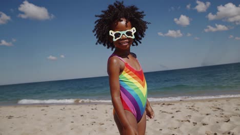 happy young girl with curly hair wearing colorful rainbow swimsuit and sunglasses enjoying summer vacation on a sunny beach with ocean waves and blue sky in background
