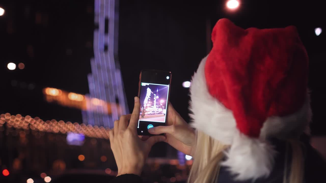 산타클로스 모자를 입은 여성이 스마트폰으로 나이트 시티의 사진을 찍습니다.