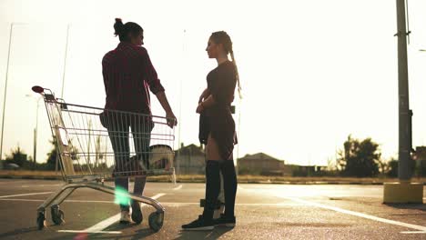 una joven atractiva con calcetines largos sostiene una patineta y habla con su novia que está sentada en el carrito de compras en el estacionamiento cerca del centro comercial durante la puesta de sol