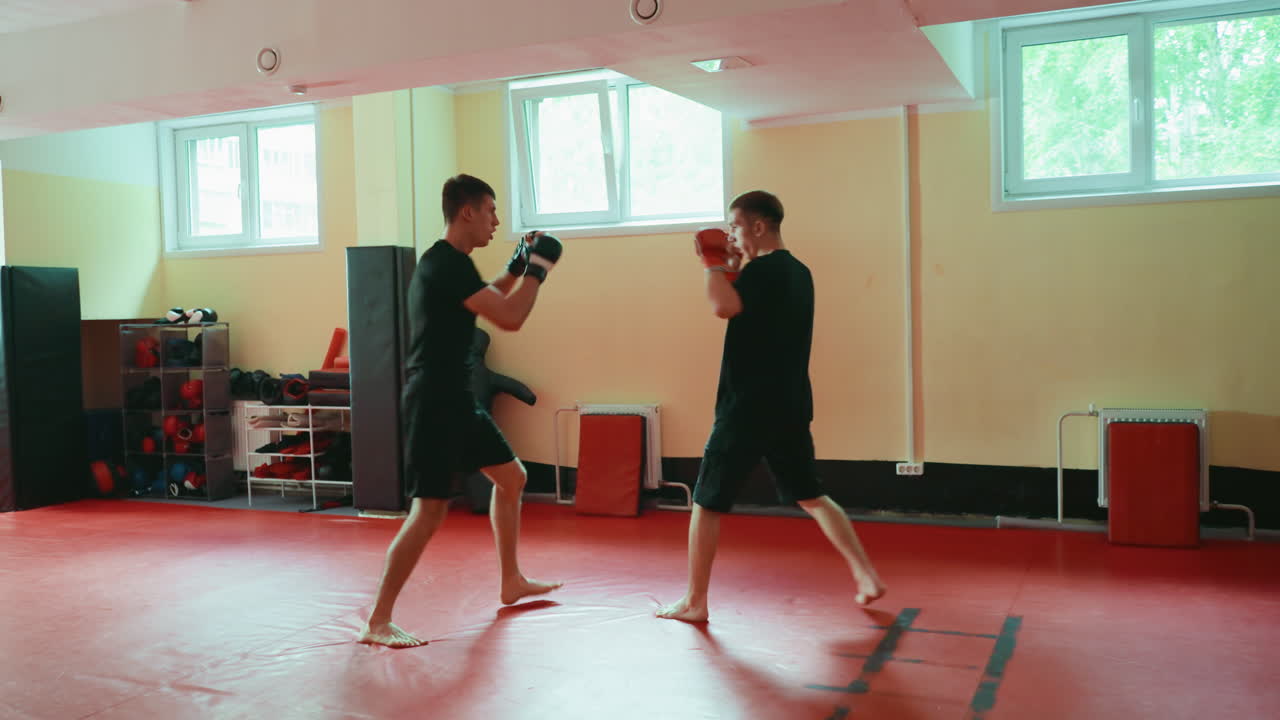 Two male fighters facing each other in martial arts gym preparing for sparring practice with boxing gloves on red mat floor under daylight from windows with training equipment visible in background