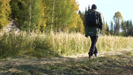 Hiking-man-with-backpack-passes-an-old-barn-and-grain-field-on-natural-walking-trail,-yellow-autumn-colors