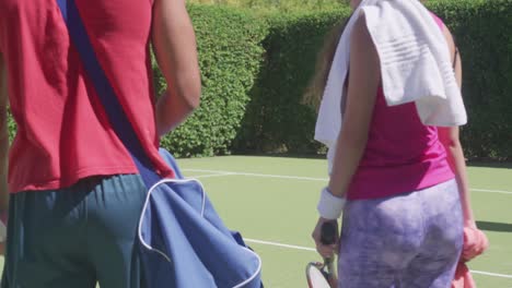 Happy-biracial-couple-walking-with-bags-and-tennis-rackets-in-garden-on-sunny-day