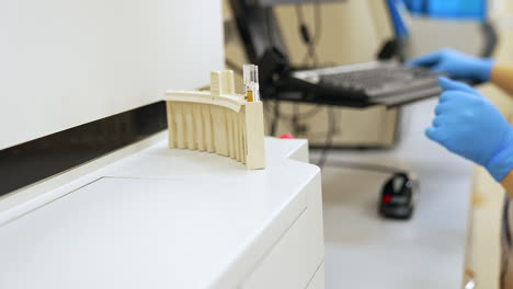a plastic stand with two test tubes inside. a laboratory professional sitting at the computer wearing blue gloves at backdrop in blur.
