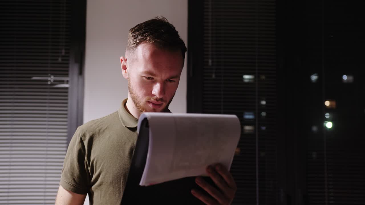 Young guy investigator working at night in the office in the department tired looking for papers looking through documents traces of criminal professionally looking at the evidence standing at the window of the night city