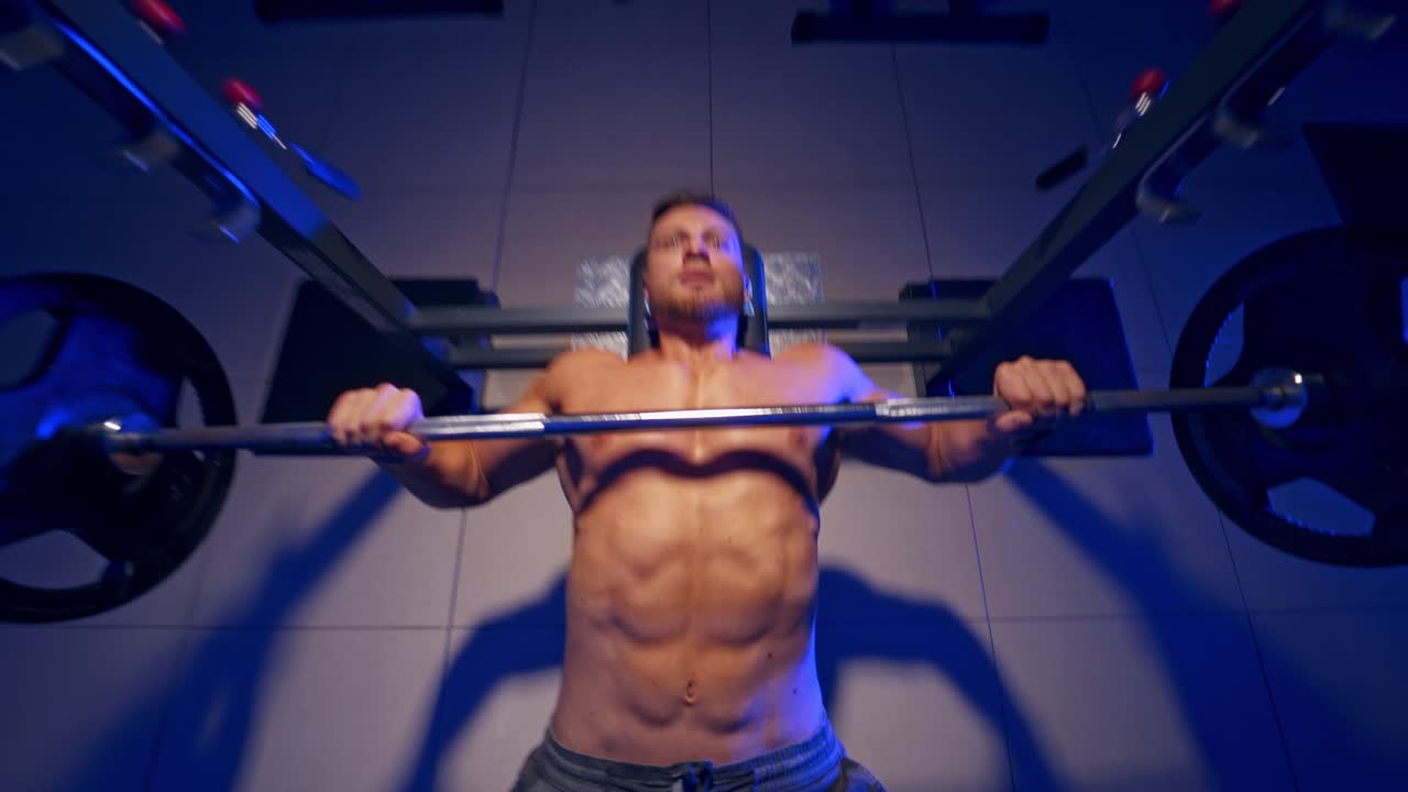 Bodybuilder trainer man doing bench press workout in gym. Video from the top. Blue light filter. Naked torso. Strong abs.
