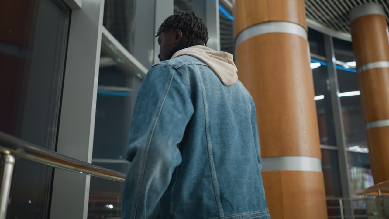 Back view of man wearing denim jacket over hoodie walking indoors through brightly lit modern building at night with large windows and architectural elements reflecting city lights and evening atmosphere