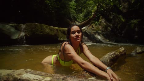 A-young-woman-in-a-sexy-bikini-kneel-on-a-log-with-a-cascading-waterfall-in-the-background