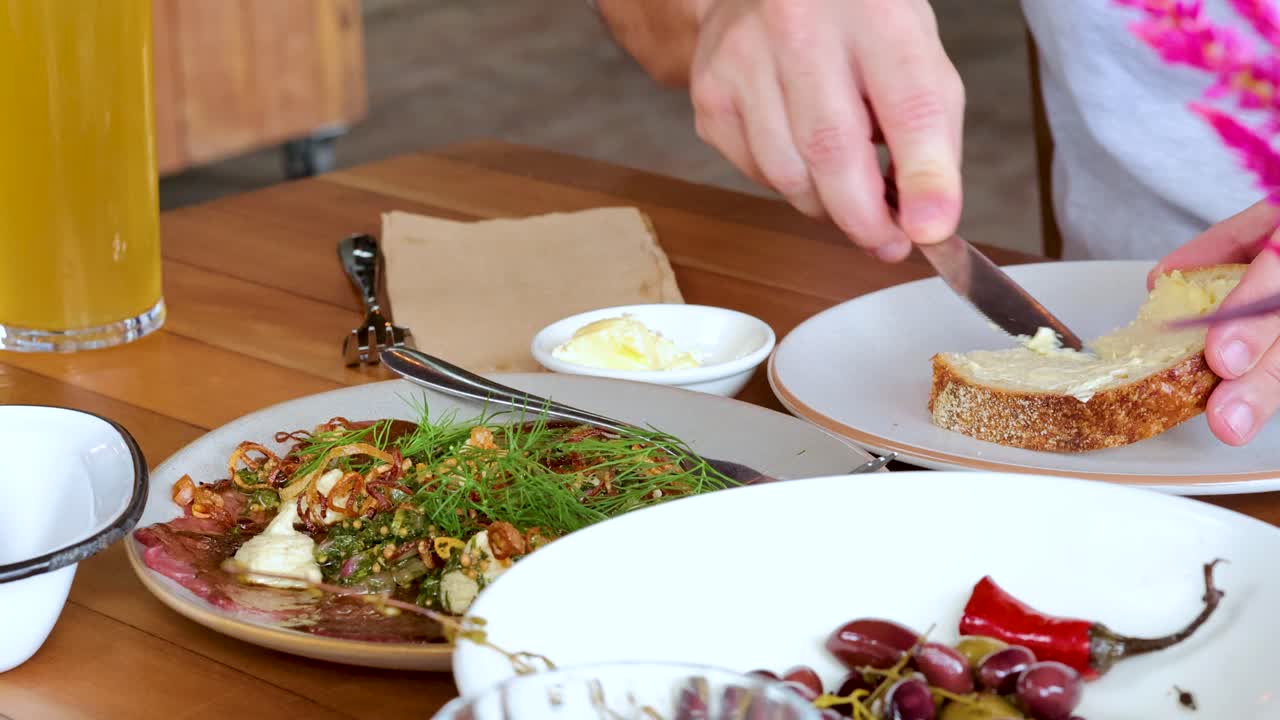 A person spreads butter on a slice of bread at a well-lit breakfast table with fresh juice, salad, cherries, and rustic decor. Static camera, natural light