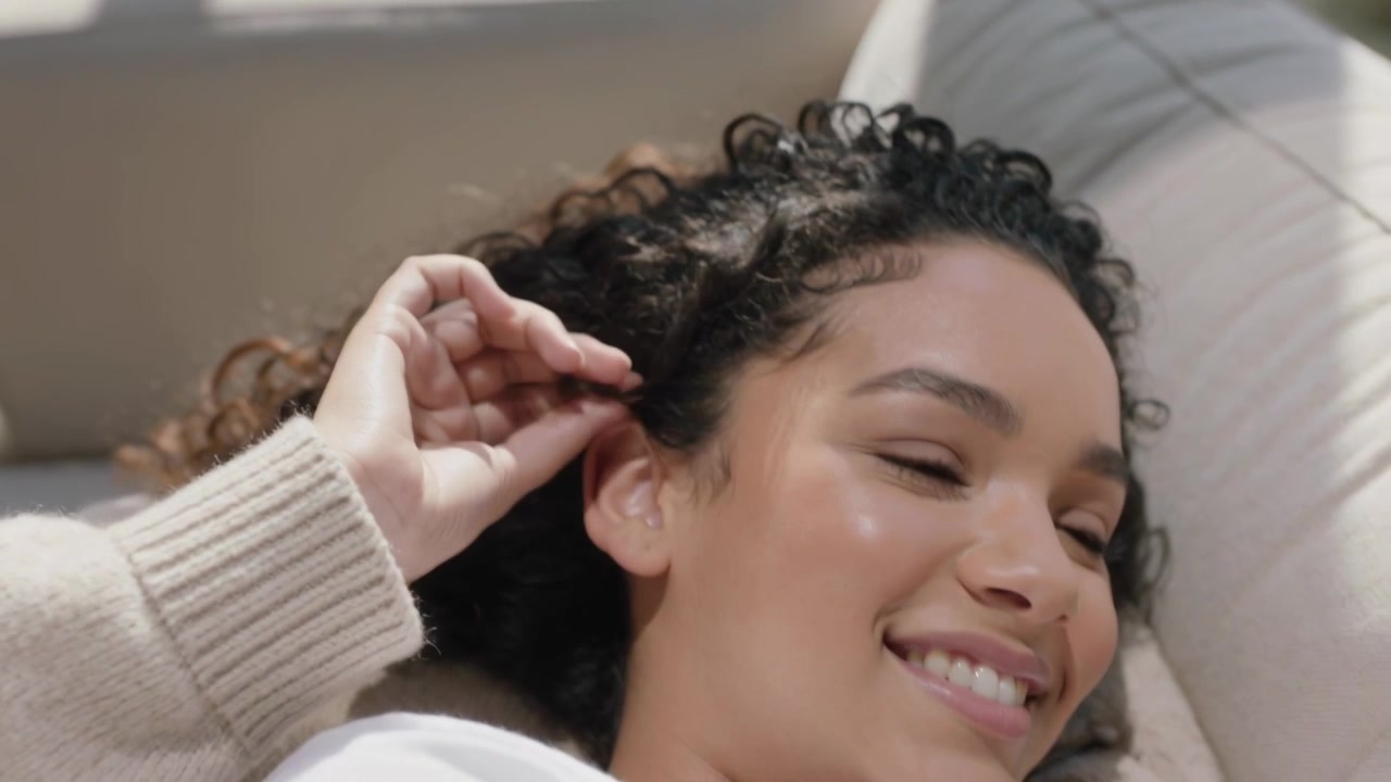 Una donna che si rilassa sul divano, si attorciglia un ricciolo di capelli e lo sistema vicino all'orecchio, mostrando la manica del maglione.