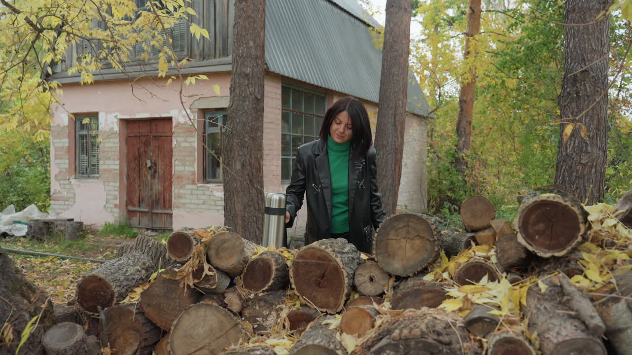 black woman in leather jacket leaning against stacked logs outside rustic cottage during autumn, holding thermos with warm green sweater, contemplative smile and relaxed pose, pine trunks and yellow