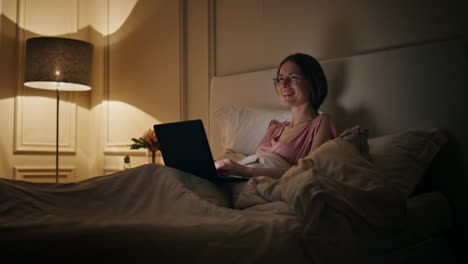 chill woman browsing laptop at night. cheerful freelancer working late in bed