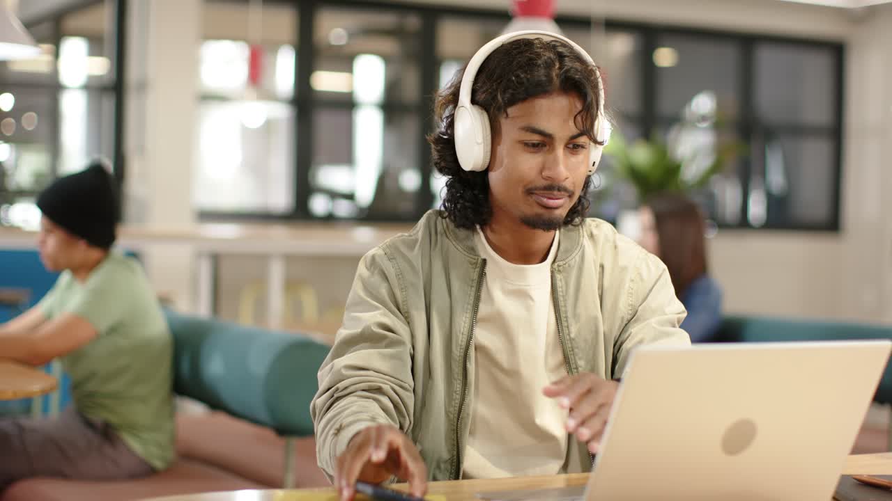 Afroamerikansk mand scroller på smartphone, skifter til bærbar computer, taster, arbejder med headset på café
