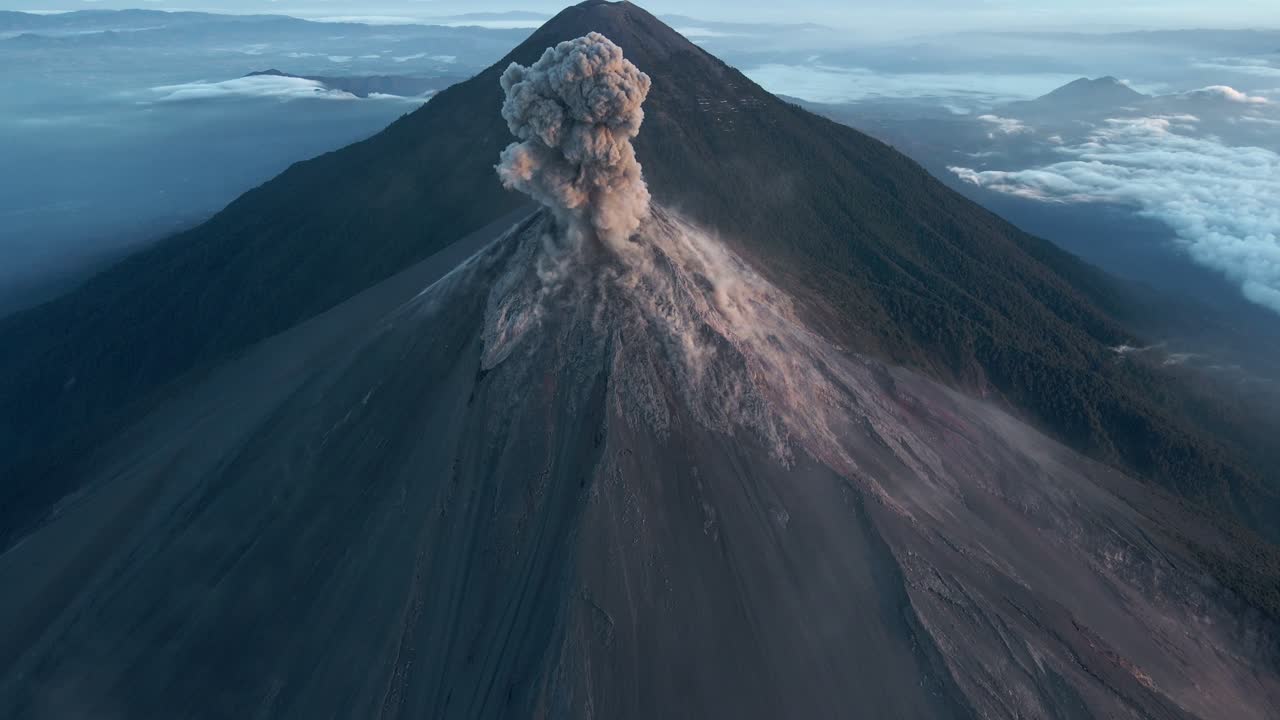 empujón aéreo cinematográfico de la erupción masiva del volcán fuego con una columna de ceniza dramáticamente ascendente y el volcán acatenango con campamentos visibles en el fondo