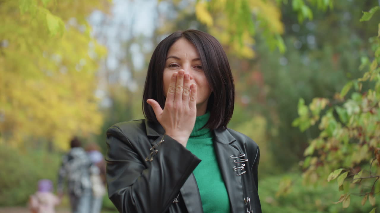 Černoška v kožené bundě posílá pusu při procházce listnatým podzimním parkem, má na sobě zelený rolák a zapletené vlasy, hravý úsměv, ruka natažená dopředu v nabízejícím gestu, ležérní rockový styl