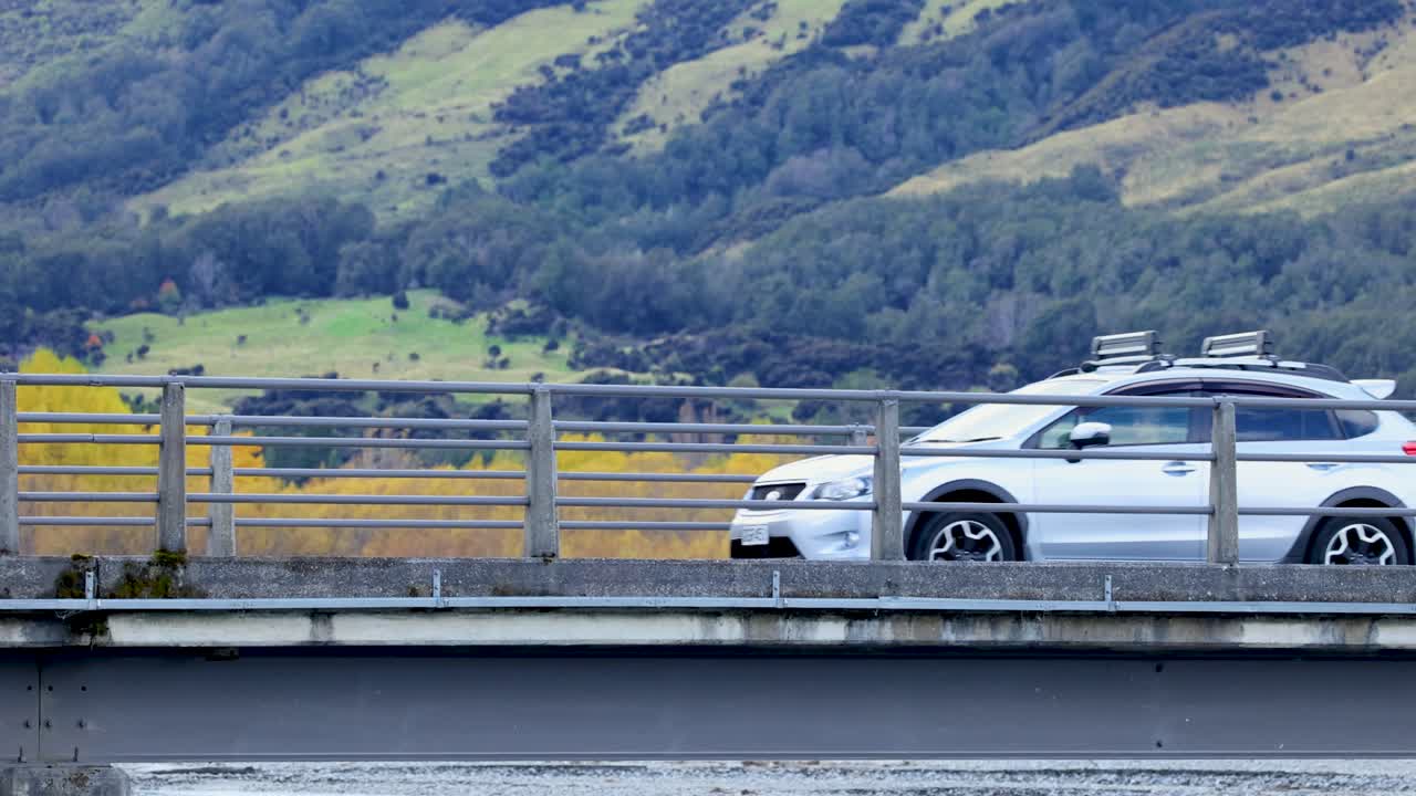 Silver SUV drives across bridge with scenic hills, steady camera, natural daylight, wide shot