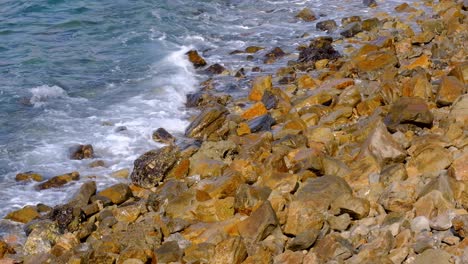Ocean-waves-rolling-in-and-crashing-onto-a-remote,-rugged-and-golden-rocky-coastal-shoreline-in-the-wilderness-on-a-tropical-island