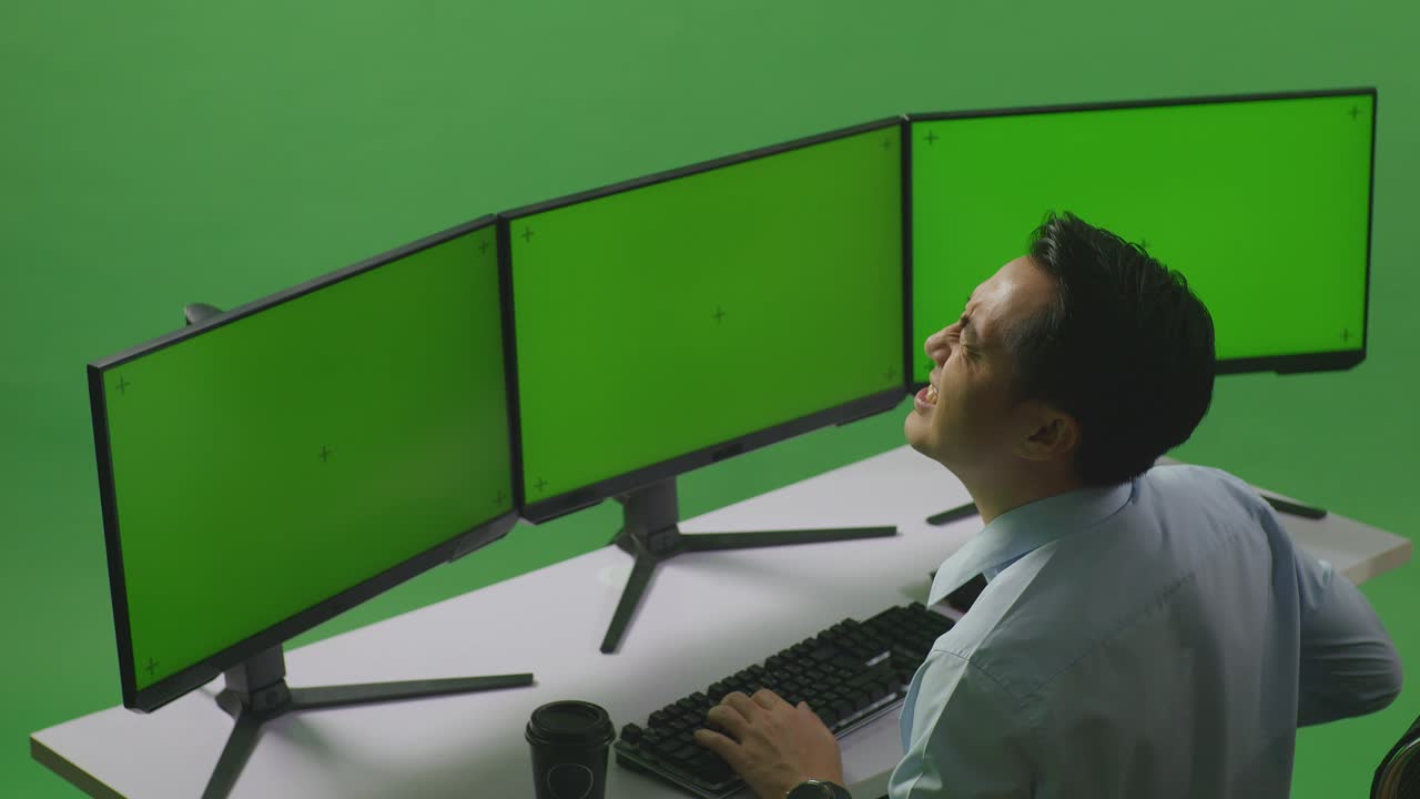 vista lateral de un hombre asiático con dolor de espalda mientras trabaja con múltiples monitores de computadora simulados en un estudio de pantalla verde