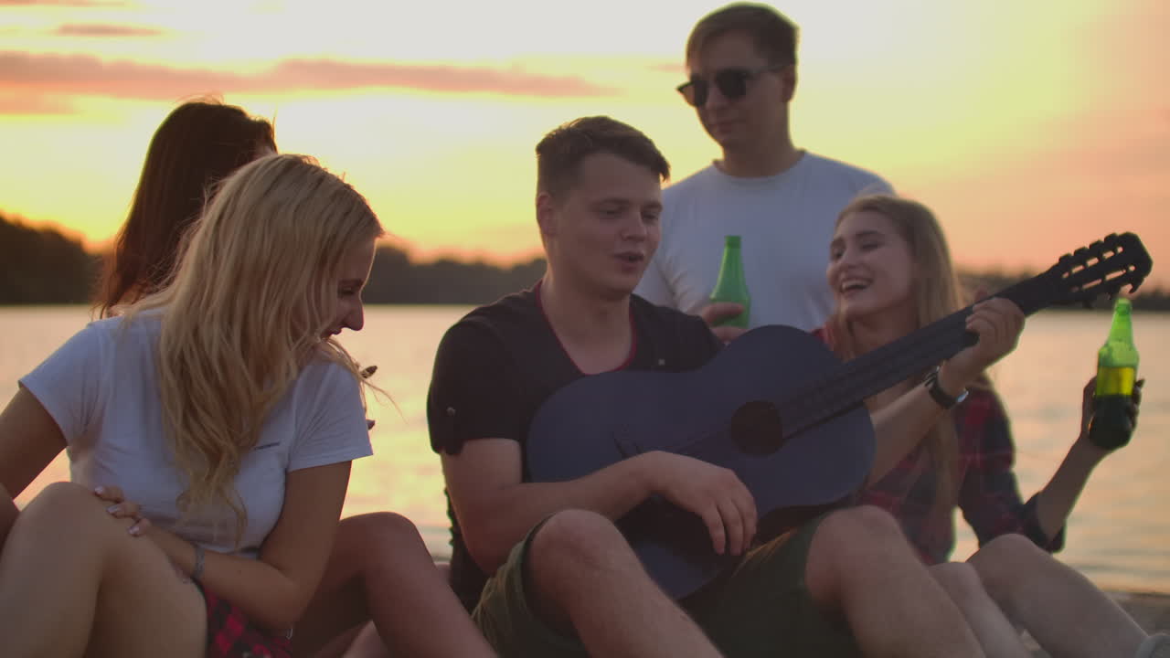 el niño está tocando la guitarra en la fiesta al aire libre con sus amigos con cerveza. esta es una noche maravillosa alrededor de la hoguera.
