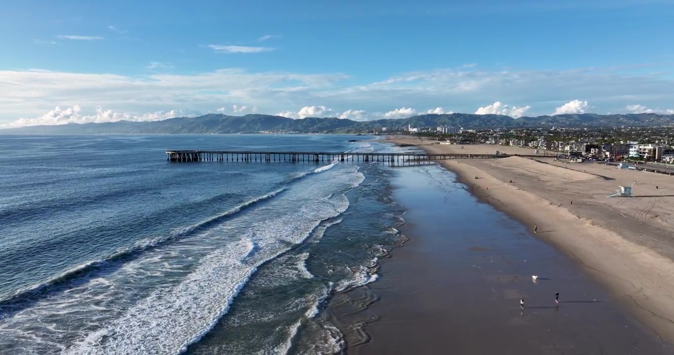 Una ripresa aerea con drone ad alta quota, cinematografica e grandangolare del molo di Santa Monica, che si allontana lentamente sopra la spiaggia di Venice e la costa del Pacifico a Los Angeles, California
