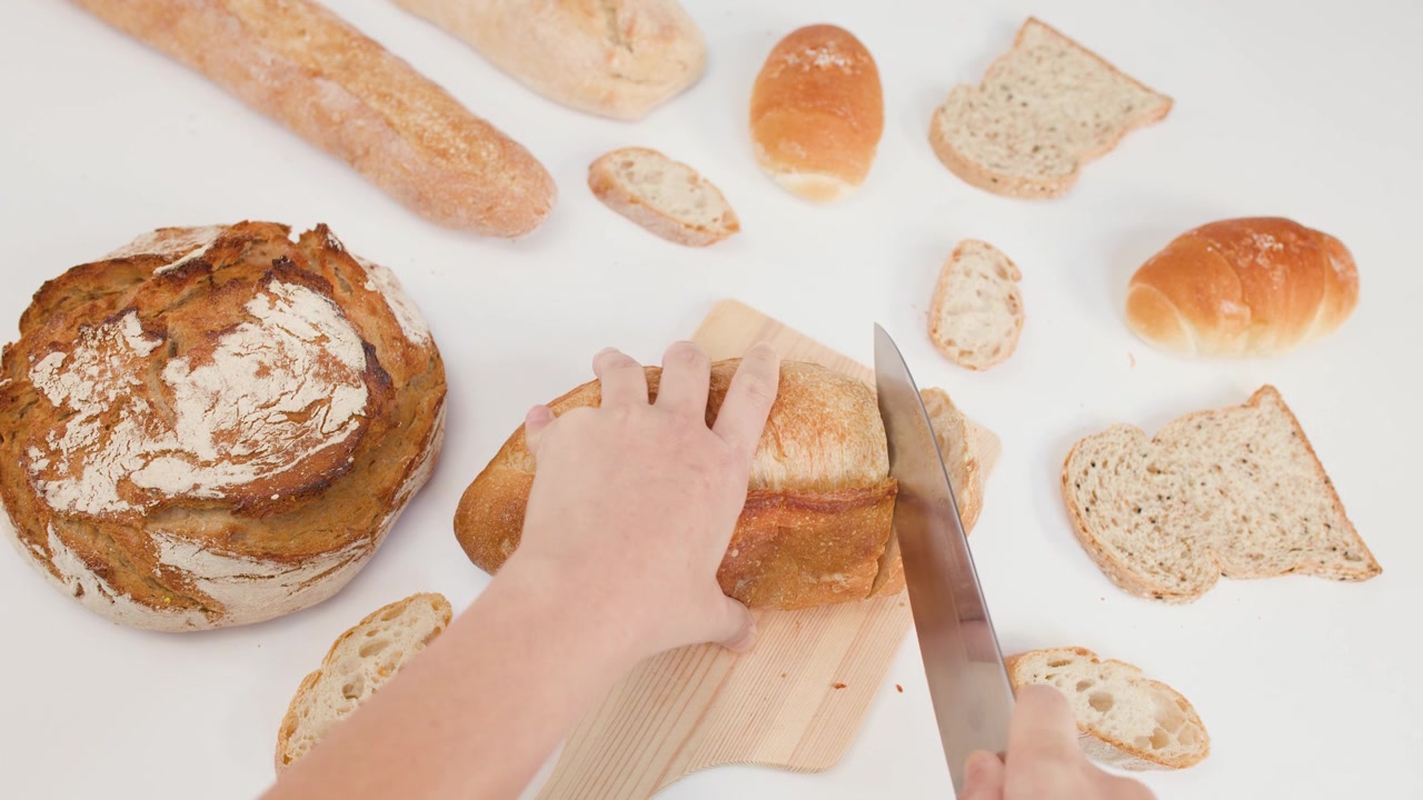 Inquadratura dall'alto di mani che usano un coltello per affettare pane croccante su un tagliere di legno.