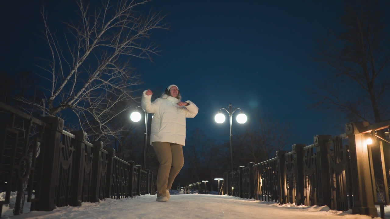 Young girl dancing along walkway decorated with warm lights and glowing lampposts under bare winter trees and crisp night sky wearing white puffer coat and headphones with expressive joyful movement