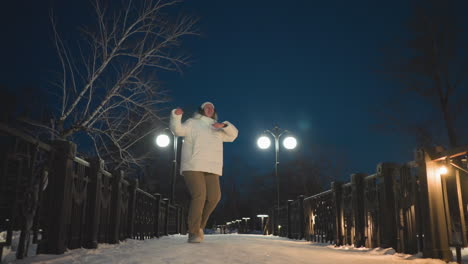 young girl dancing along walkway decorated with warm lights and glowing lampposts under bare winter trees and crisp night sky wearing white puffer coat and headphones with expressive joyful movement