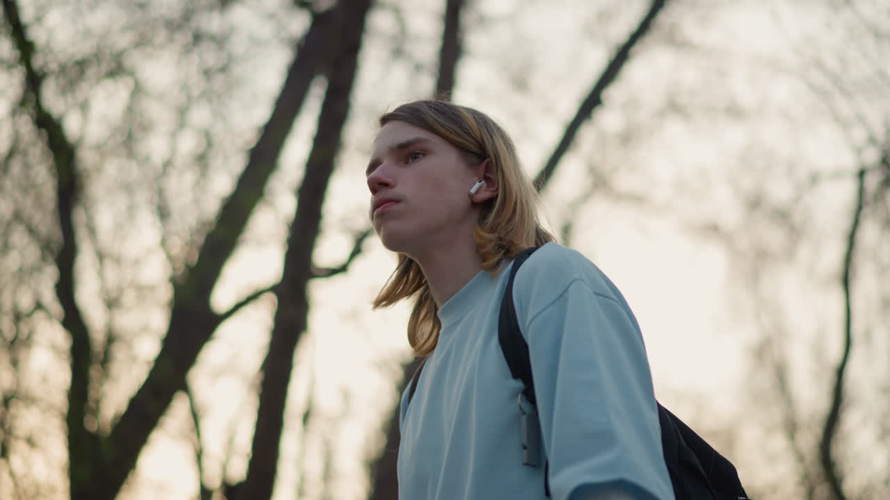 estudiante blanco caminando por un parque al amanecer con una taza de café y una mochila, expresión contemplativa, silueta de árboles desnudos, brisa fresca, paso lento y constante, luz de fondo cinematográfica y paleta de colores neutros