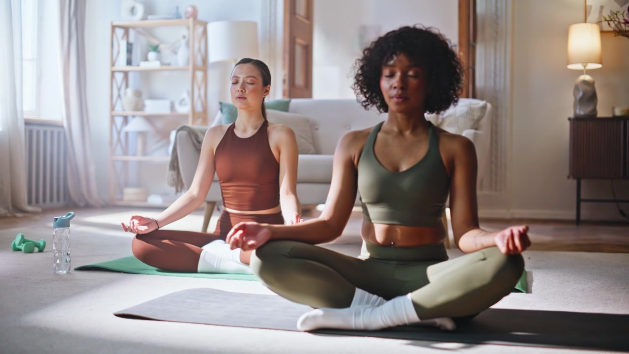 Kvinnor med olika bakgrund mediterar i lotusställning hemma. Lugn yogameditation tillsammans på morgonen