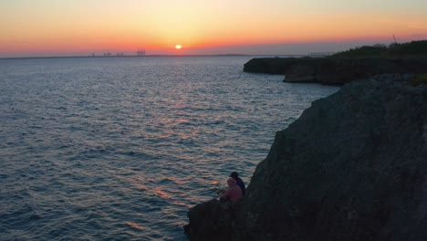 Vista-Aérea-De-Dos-Pescadores-Dominicanos-Saludando-Sentados-En-Un-Acantilado-Con-Puesta-De-Sol-En-El-Fondo,-Pedernales