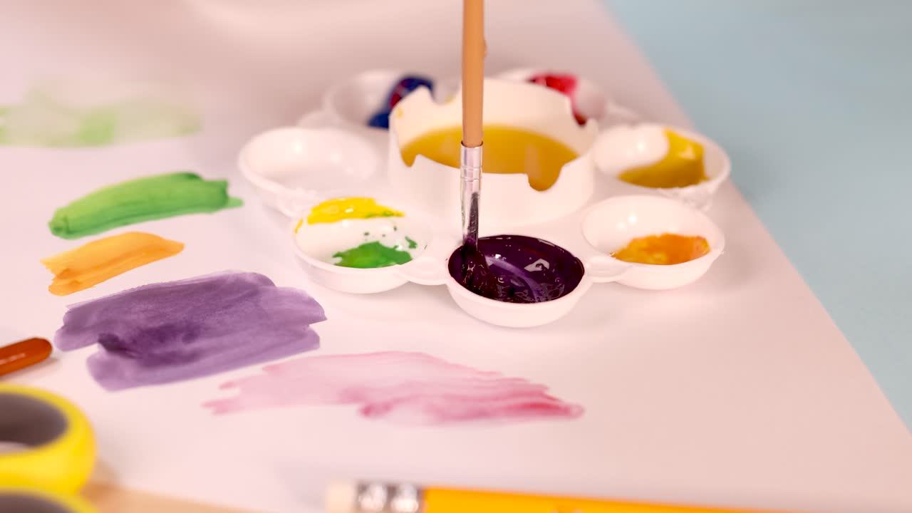 Artist hand dips brush into purple paint on palette, bright lighting, close-up, creative workspace