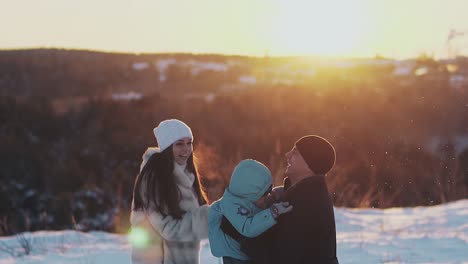 Eltern-Spielen-Im-Winter-Und-Werfen-Ihr-Kind-Gegen-Den-Orangefarbenen-Sonnenuntergang