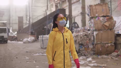 Ein-Selbstbewusstes-Mädchen-Mit-Dreadlocks-In-Schützender-Arbeitskleidung,-Gelber-Jacke,-Brille-Und-Maske,-Die-An-Der-Recyclingfabrik-Vorbeigeht