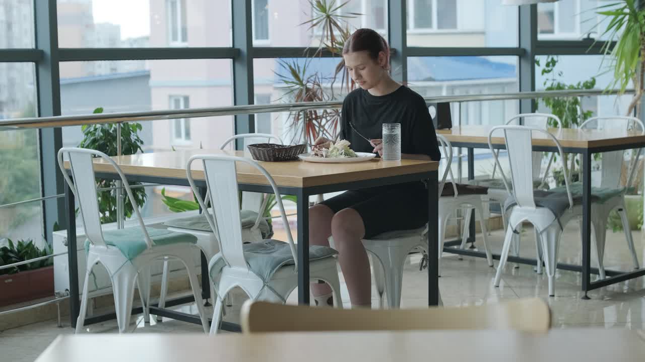 mujer joven comiendo una ensalada fresca en una mesa de madera en un restaurante moderno con grandes ventanas, luz natural y vistas urbanas. escena de comedor relajada