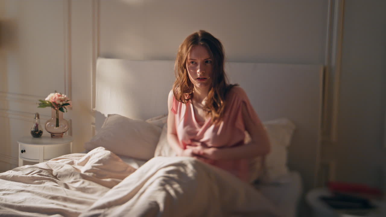 침대에 누워있는 여성은 배통을 느니다. 좌절된 깨어있는 소녀는 배를 만지고 있습니다.