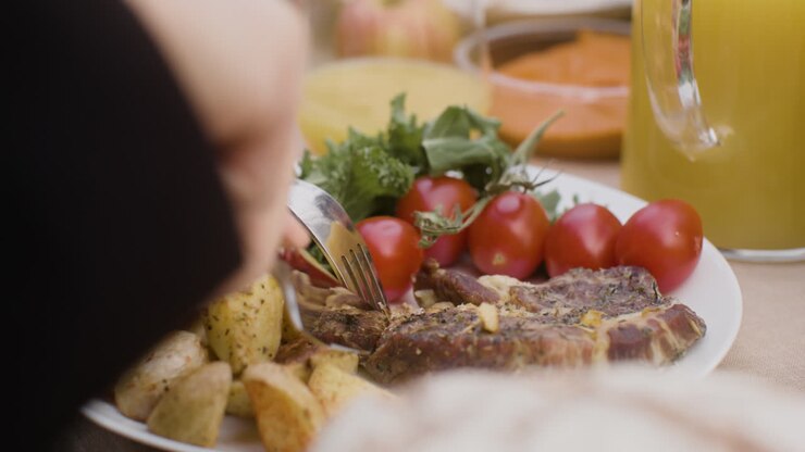 vista de cerca de un hombre cortando a mano un filete de carne de un plato con verduras y patatas durante una fiesta al aire libre en el parque