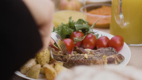 Vista-De-Cerca-De-Un-Hombre-Cortando-A-Mano-Un-Filete-De-Carne-De-Un-Plato-Con-Verduras-Y-Patatas-Durante-Una-Fiesta-Al-Aire-Libre-En-El-Parque