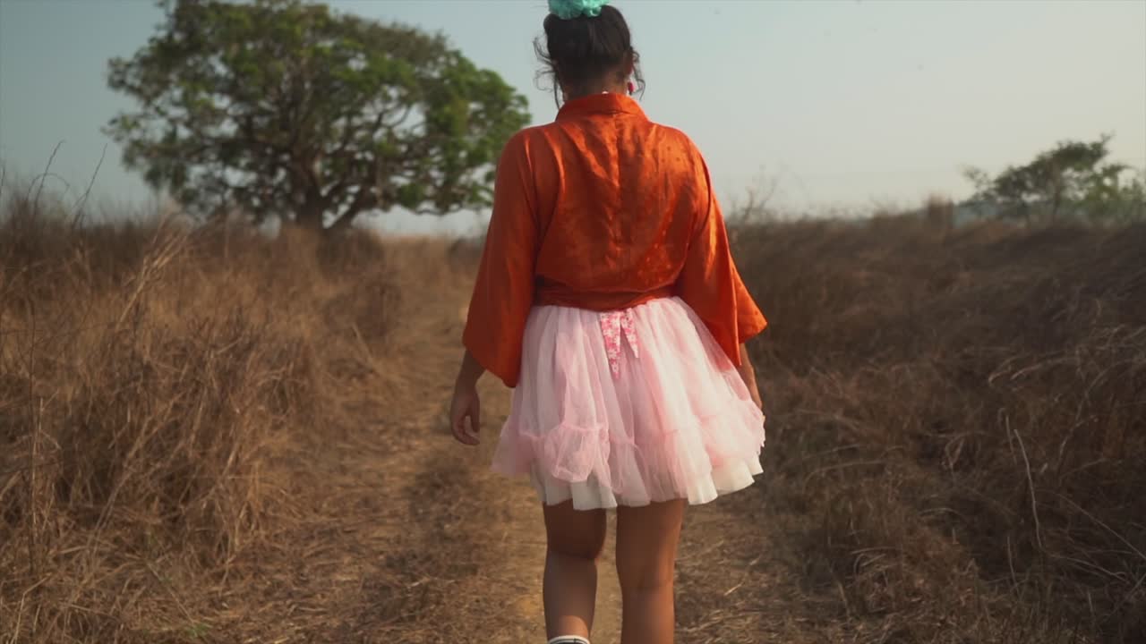 una toma en cámara lenta de la parte posterior de una mujer asiática despreocupada y saliente con un vestido de tutú rosa, estirando los brazos mientras camina por un sendero en un campo de hierba seca, india