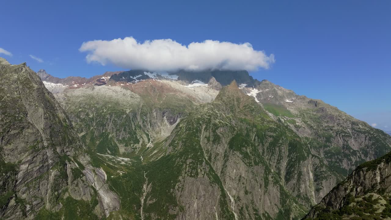 Alpine landscape featuring towering granite peaks and lush green slopes under a vivid blue sky, thick cloud formation across the mountaintops
