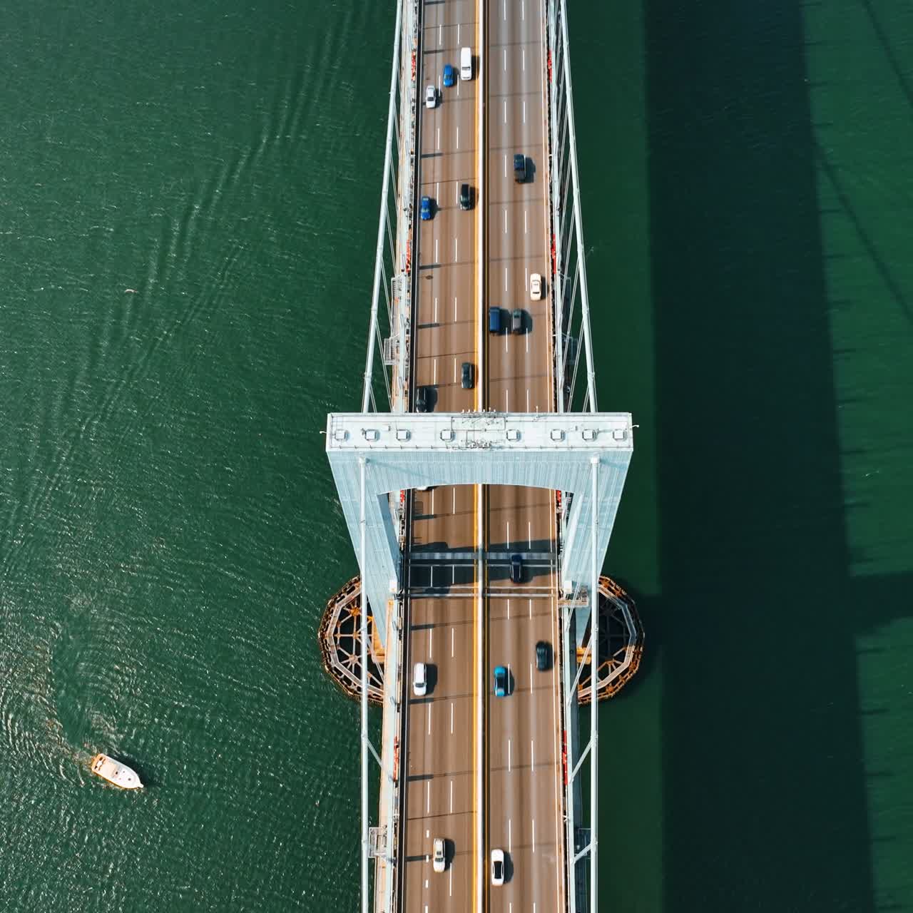 Rising above the support of suspension bridge. Numerous cars run quickly by the roads on the bridge. Yacht sails by the green water. Top view