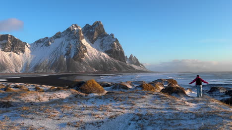 turista cargando mochila explorando la zona montañosa de vestrahorn en islandia, caminando en tierras salvajes de la costa nevada en invierno, montaña rocosa y costa en el fondo, paisaje panorámico