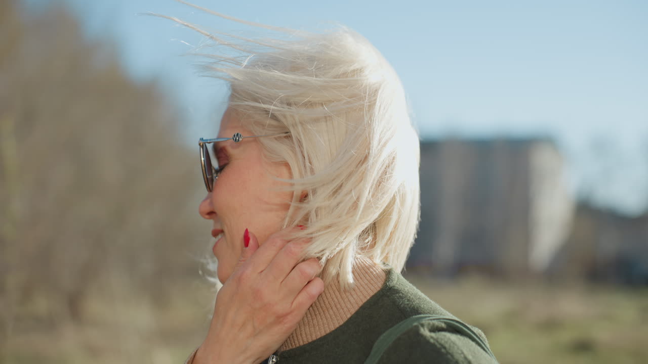 Smiling woman with blonde hair wearing sunglasses and green coat enjoying sunny day outdoors, wind blowing through hair, profile view capturing carefree moment, happiness, and relaxed lifestyle