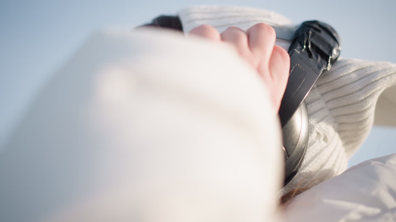 Side view of young girl wearing over ear headphones and futuristic goggles, donning knitted cap and puffer coat, smiling gently as she adjusts headset under bright winter sun, enjoying music outdoors