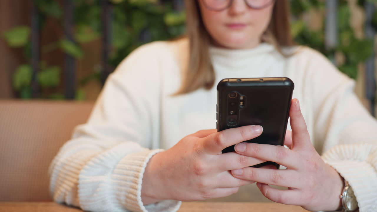 Young woman in white sweater focuses on smartphone while seated at wooden table, hands holding device as she interacts with screen, slightly blurred background featuring