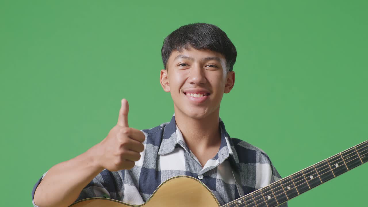 기타를 들고 있는 아시아 청소년의 근접 사진. 그린 스크린 배경에서 엄지손가락을 올려 놓고 미소 짓고 있습니다.